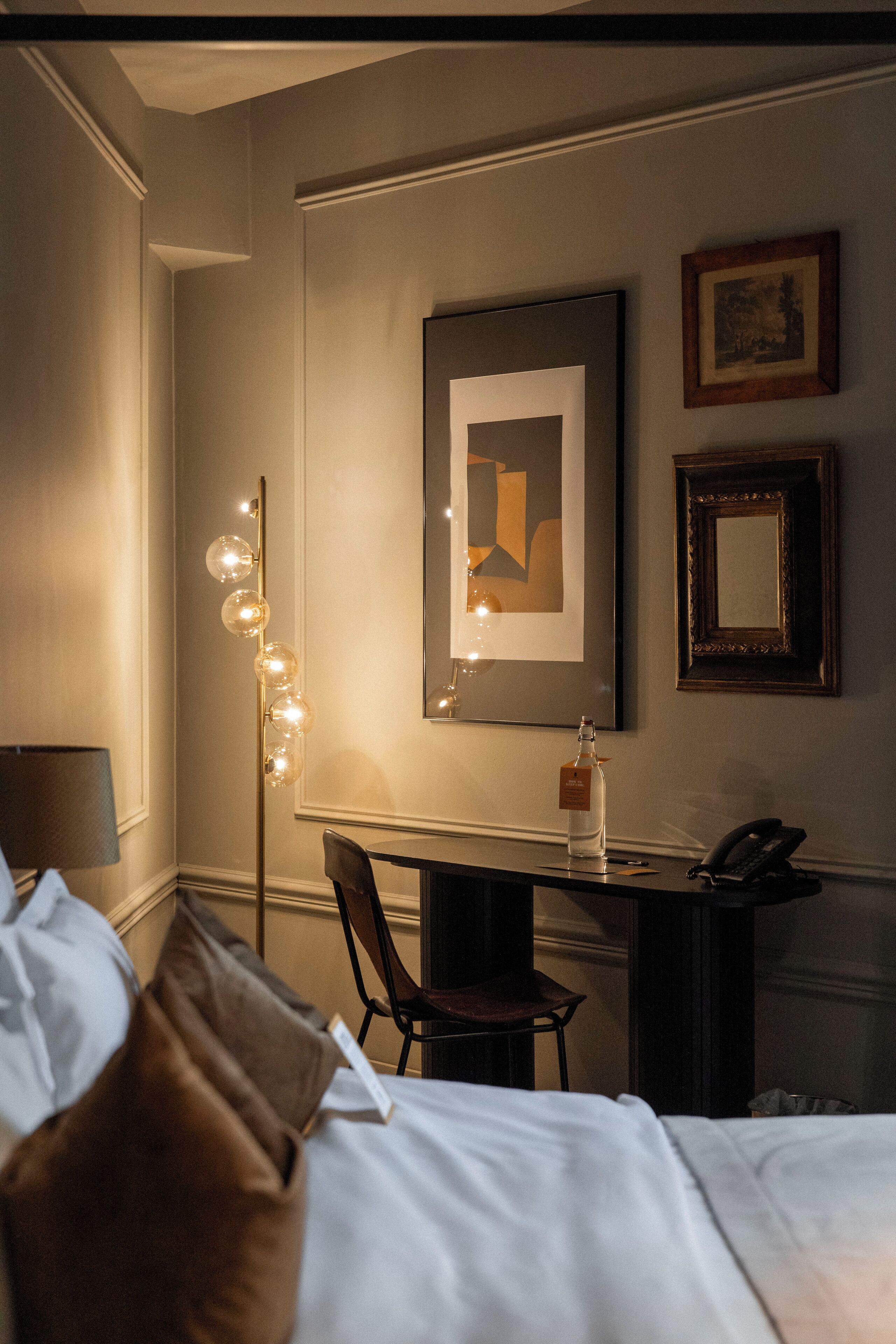 A hotel room with a bed, a desk, a chair, a lamp, and framed artwork on the wall.