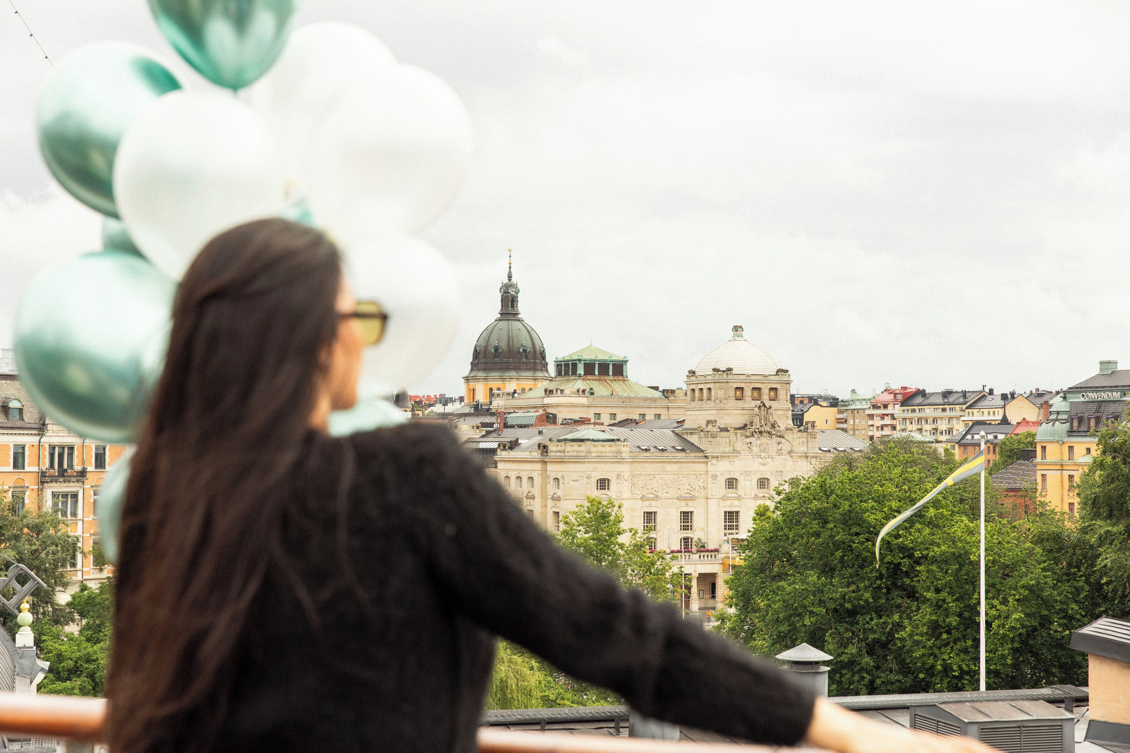 A person with balloons on a balcony overlooking a historic building with domes and classical architecture.