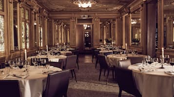 Un comedor formal con varias mesas dispuestas para una comida, grandes ventanales y detalles ornamentales en el techo.