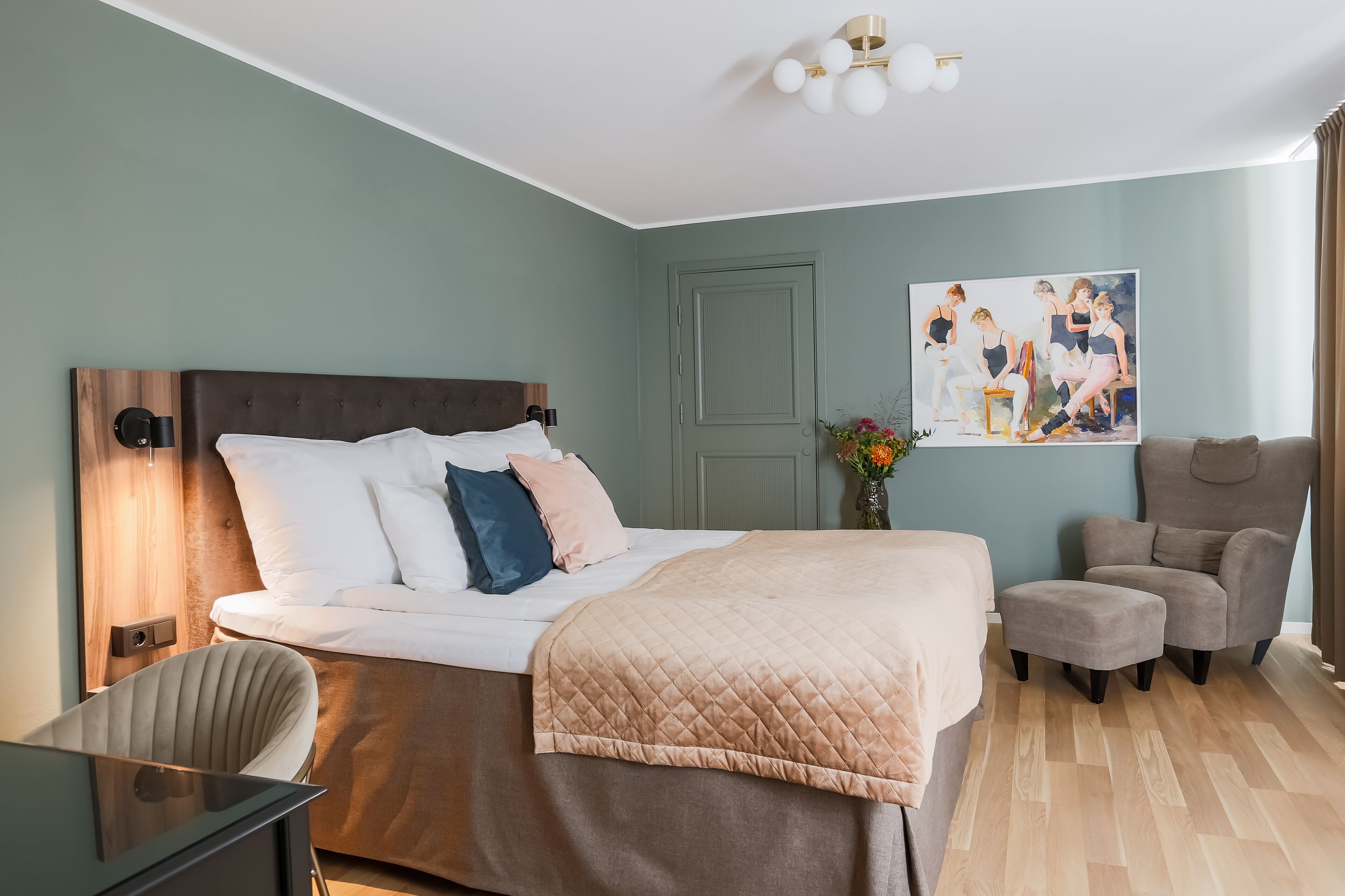 Habitación de hotel con una cama grande, un escritorio, una silla, un sillón gris y un cuadro en la pared.