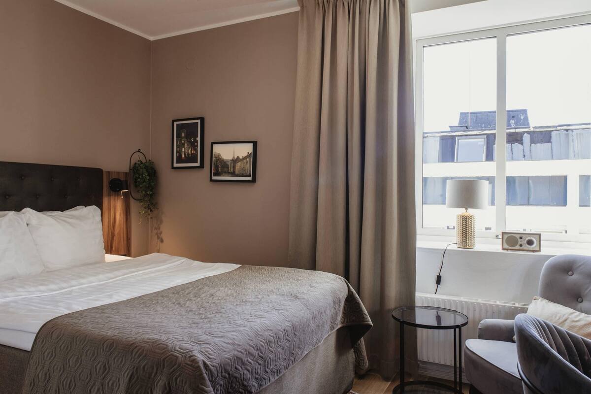 Una habitación de hotel con una cama grande, una silla, una mesita y una ventana con cortinas.