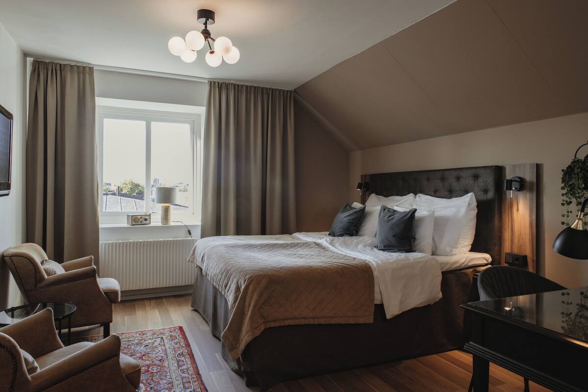 Un dormitorio con una cama grande, una zona de estar con sillas, una mesa y un ventanal con cortinas.