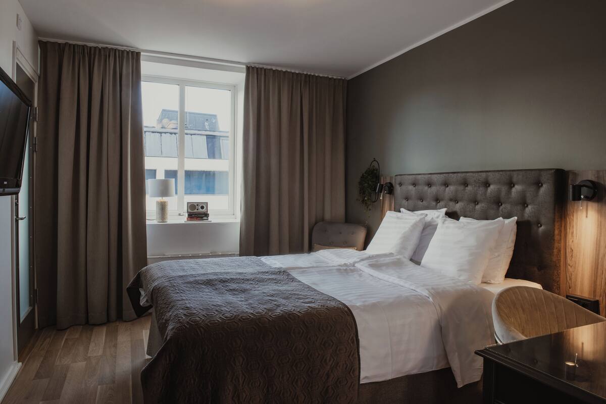 Habitación de hotel con una cama grande, una silla, una mesita de noche, un televisor y una ventana con cortinas.