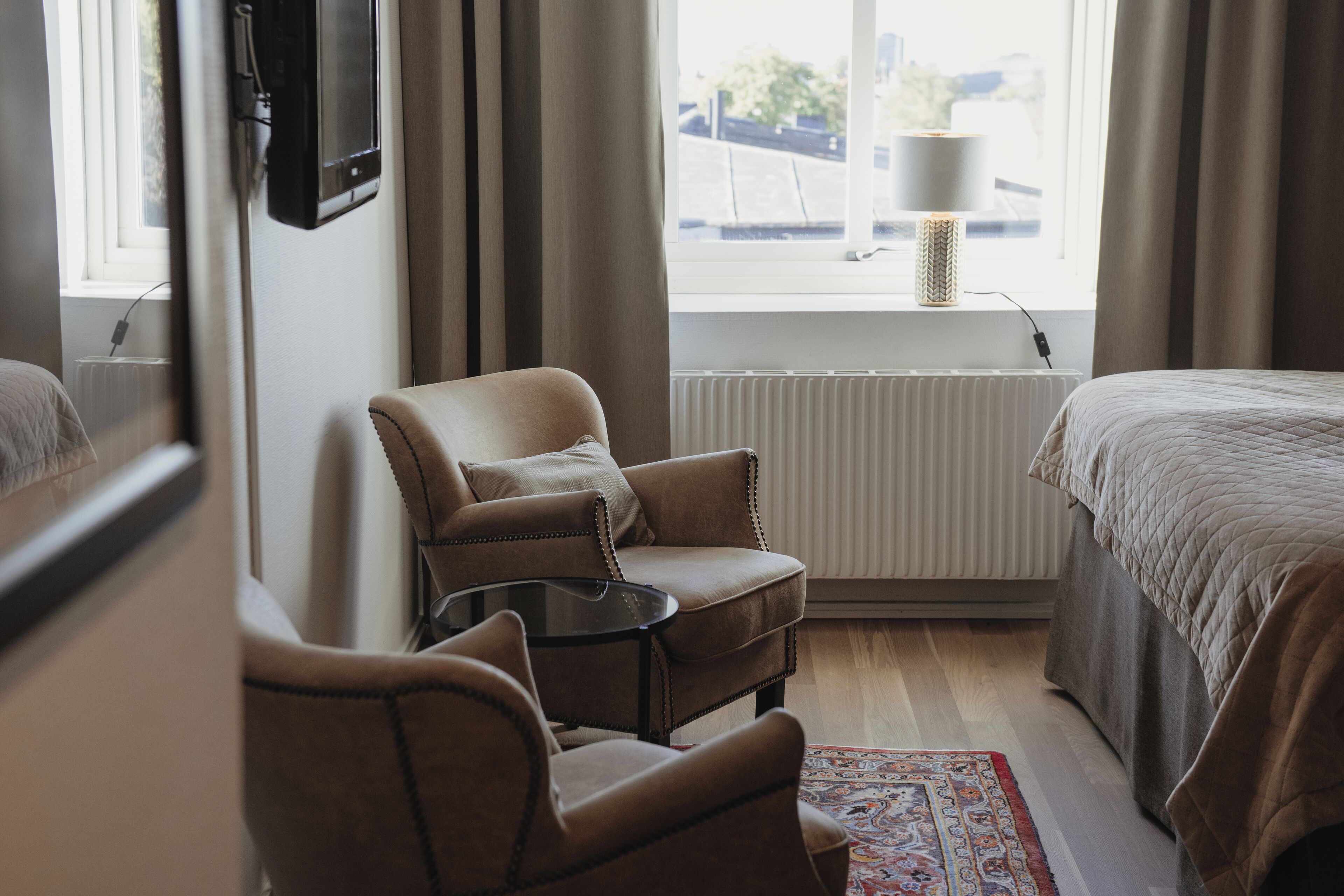 Une chambre d’hôtel avec un lit, une chaise, une petite table, un radiateur et une fenêtre avec des rideaux.