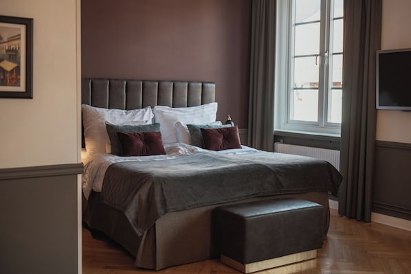 A bedroom with a large bed, a bench, a window, and a TV on the wall.