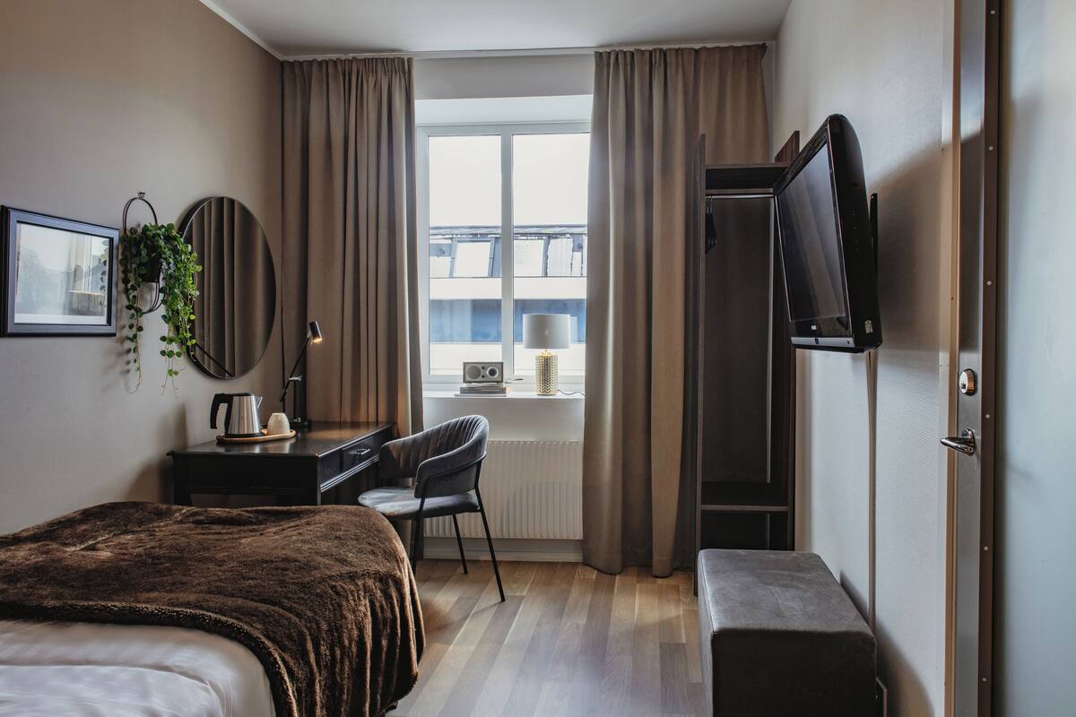 Una habitación de hotel con cama, escritorio con silla, televisor y ventana con cortinas.