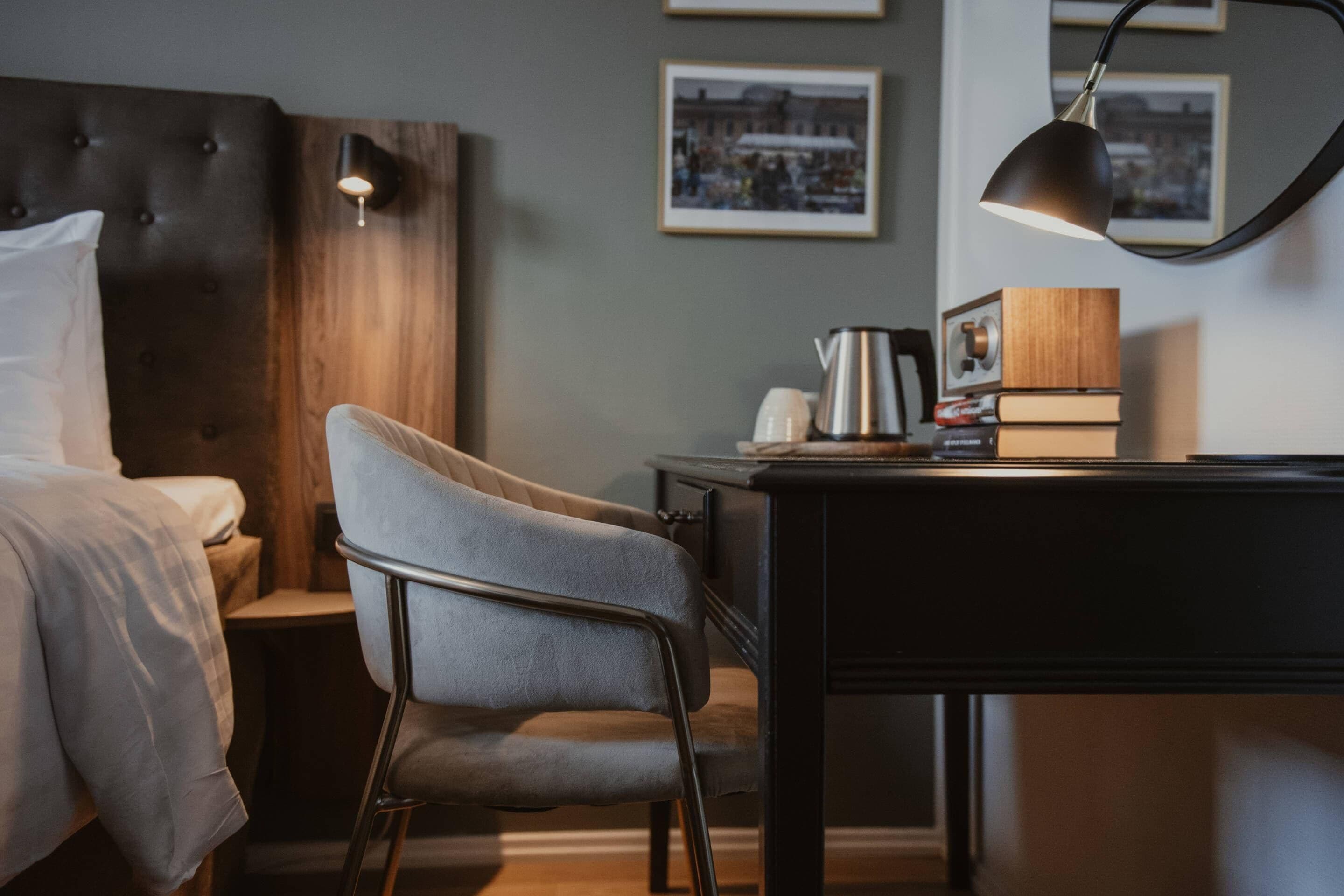 A hotel room with a bed, a chair, a desk, a lamp, and framed pictures on the wall.