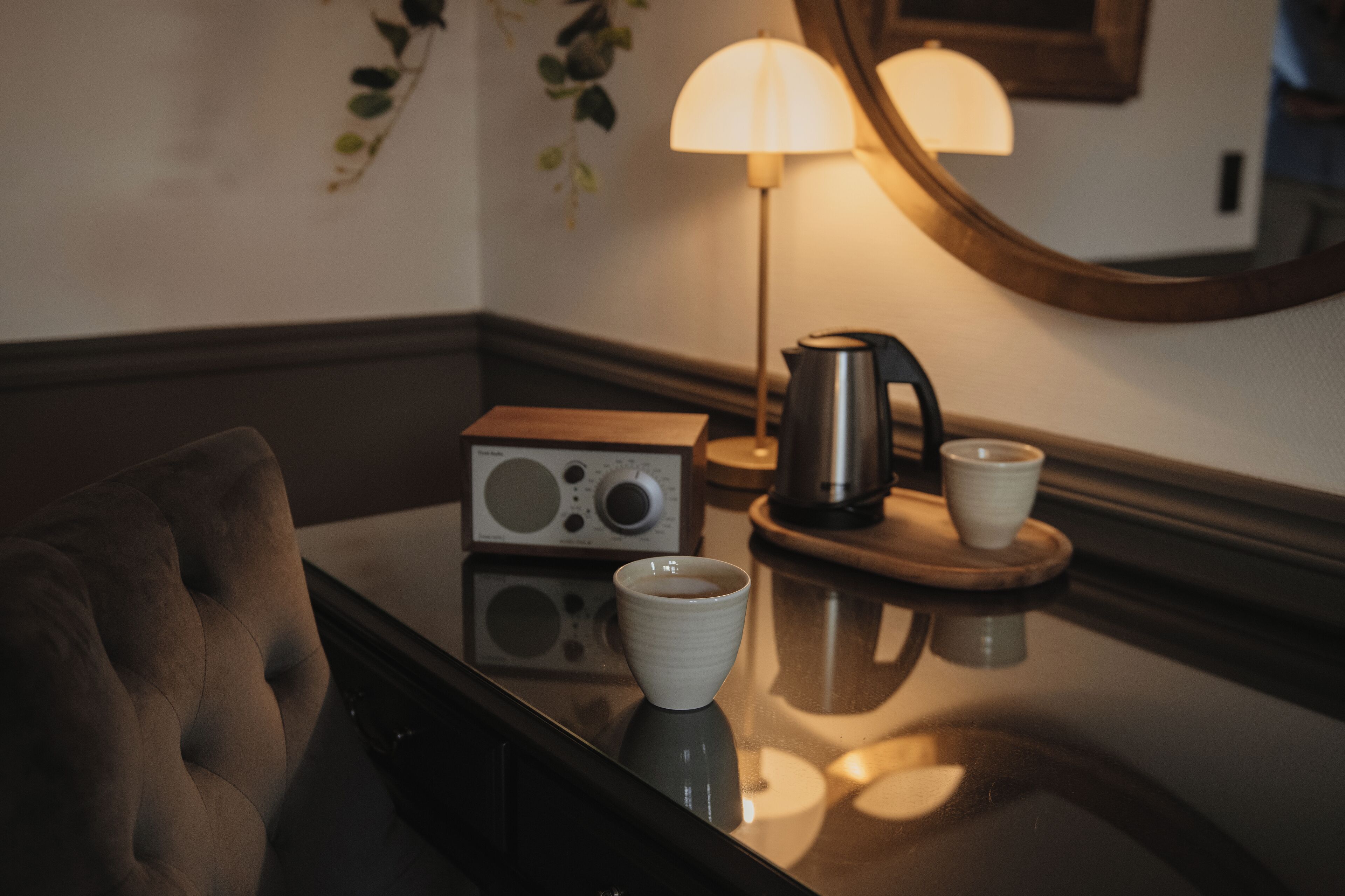 Una habitación de hotel acogedora con un sillón mullido, una cafetera, una taza y una lámpara sobre una superficie reflectante.