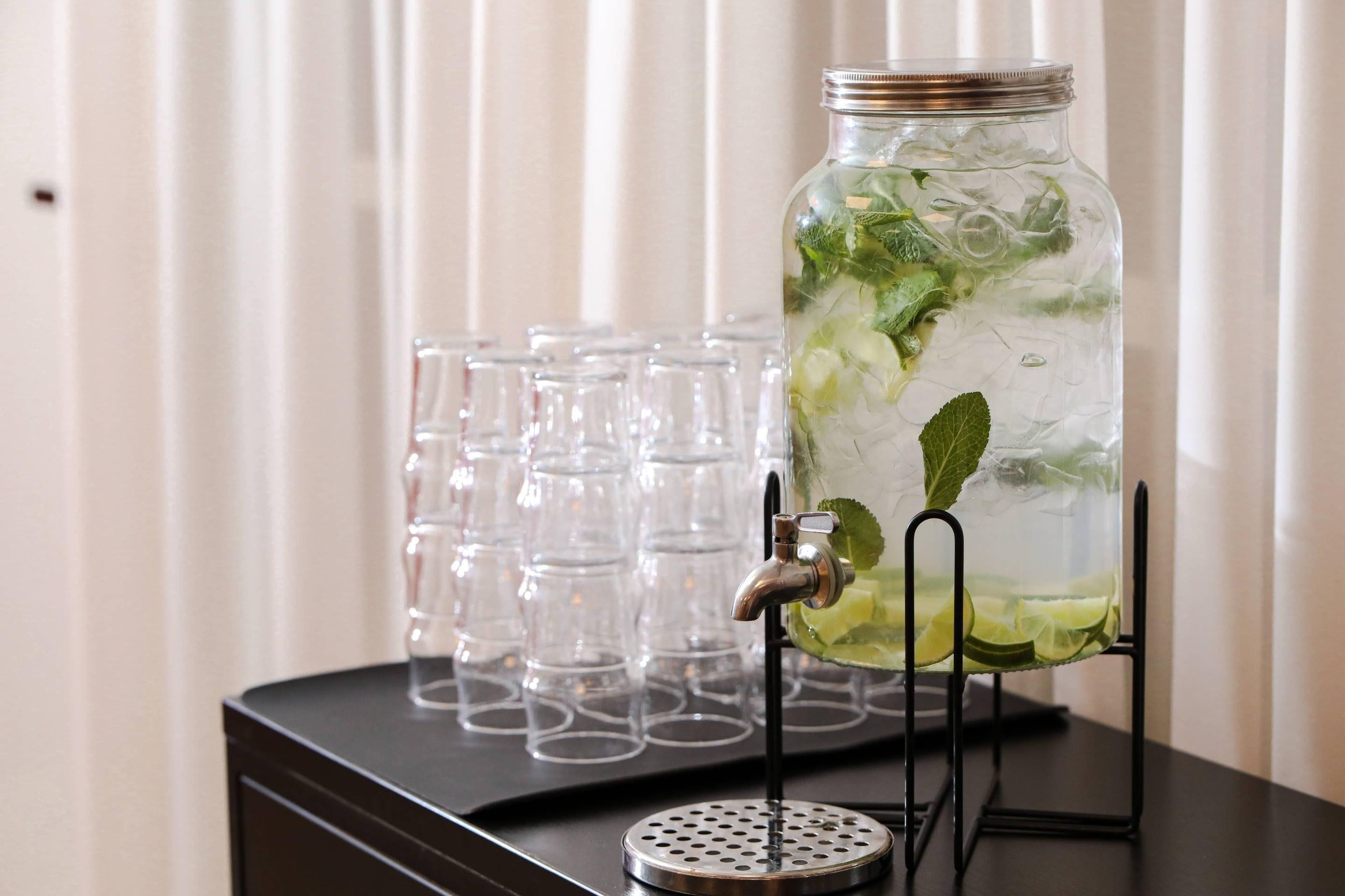 A clear glass jar with a metal tap filled with a beverage, surrounded by empty glasses on a tray.