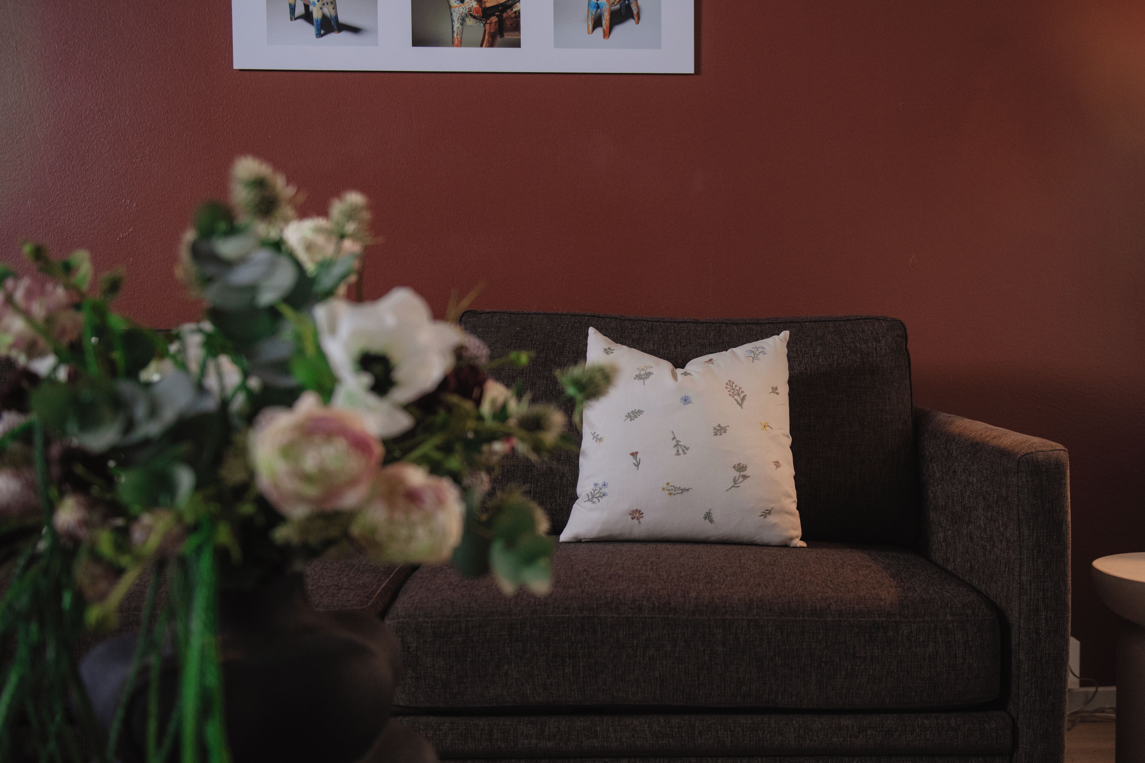 Una habitación con un sofá gris oscuro, un cojín blanco estampado con flores, y un florero con flores en primer plano.