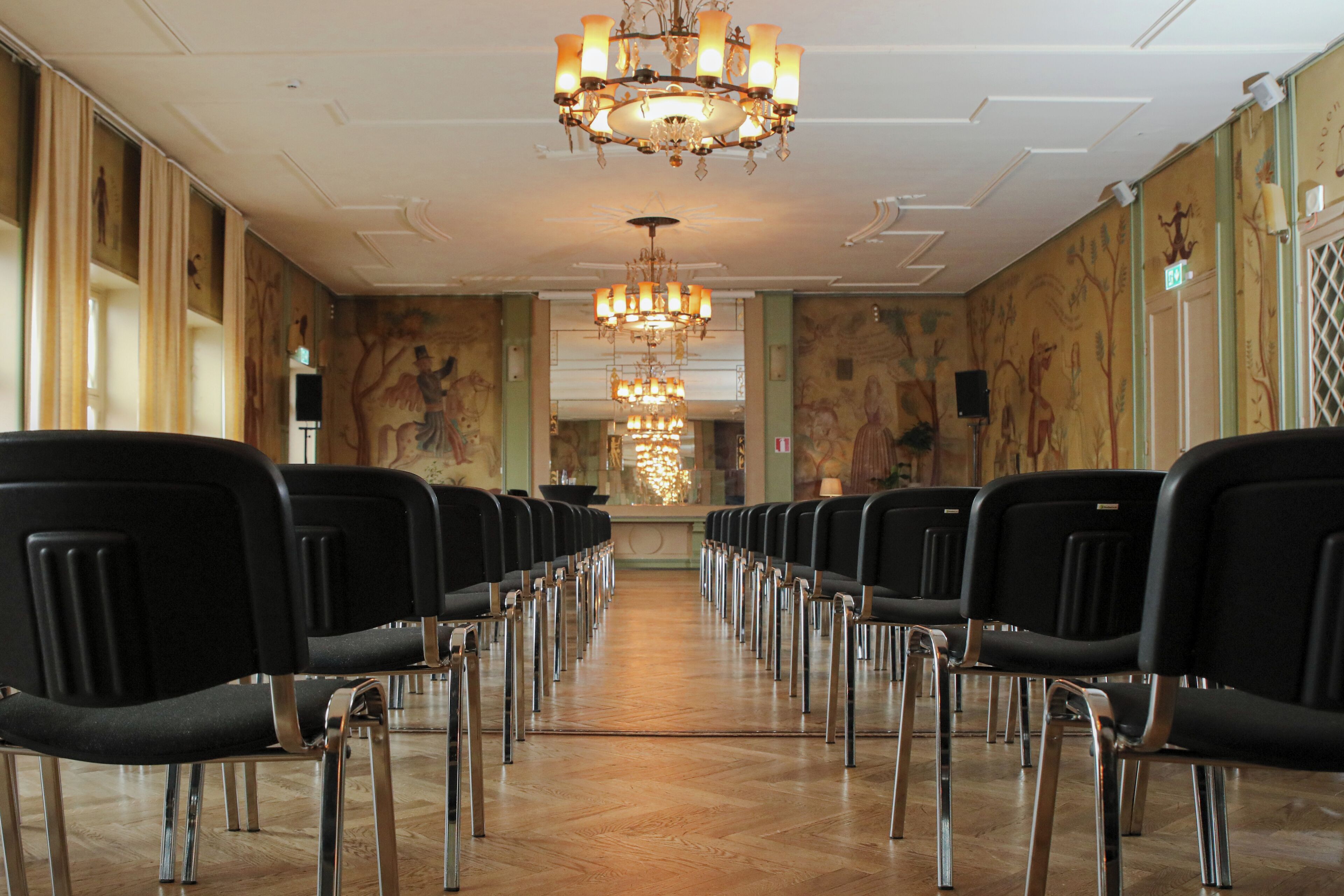 A long row of black chairs with metal legs in a room with a wooden floor and a chandelier.