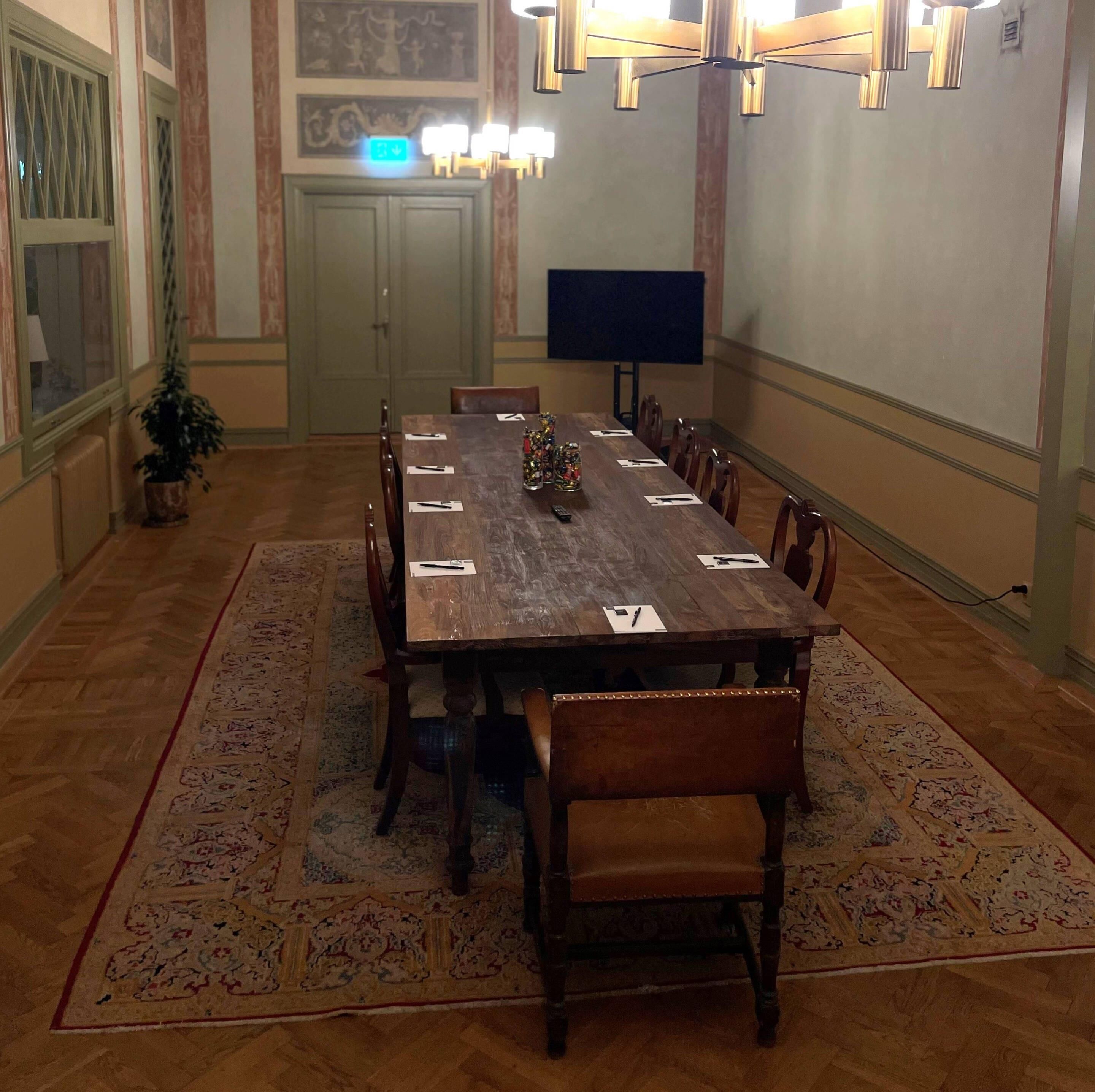 Un salón de conferencias con una larga mesa, sillas, un televisor y una planta decorativa.