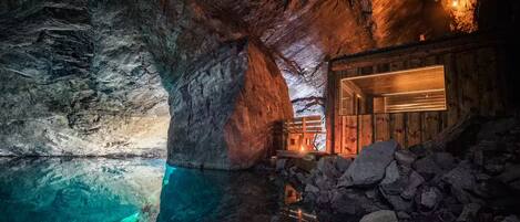 A cave interior with a wooden cabin and a clear water pool.