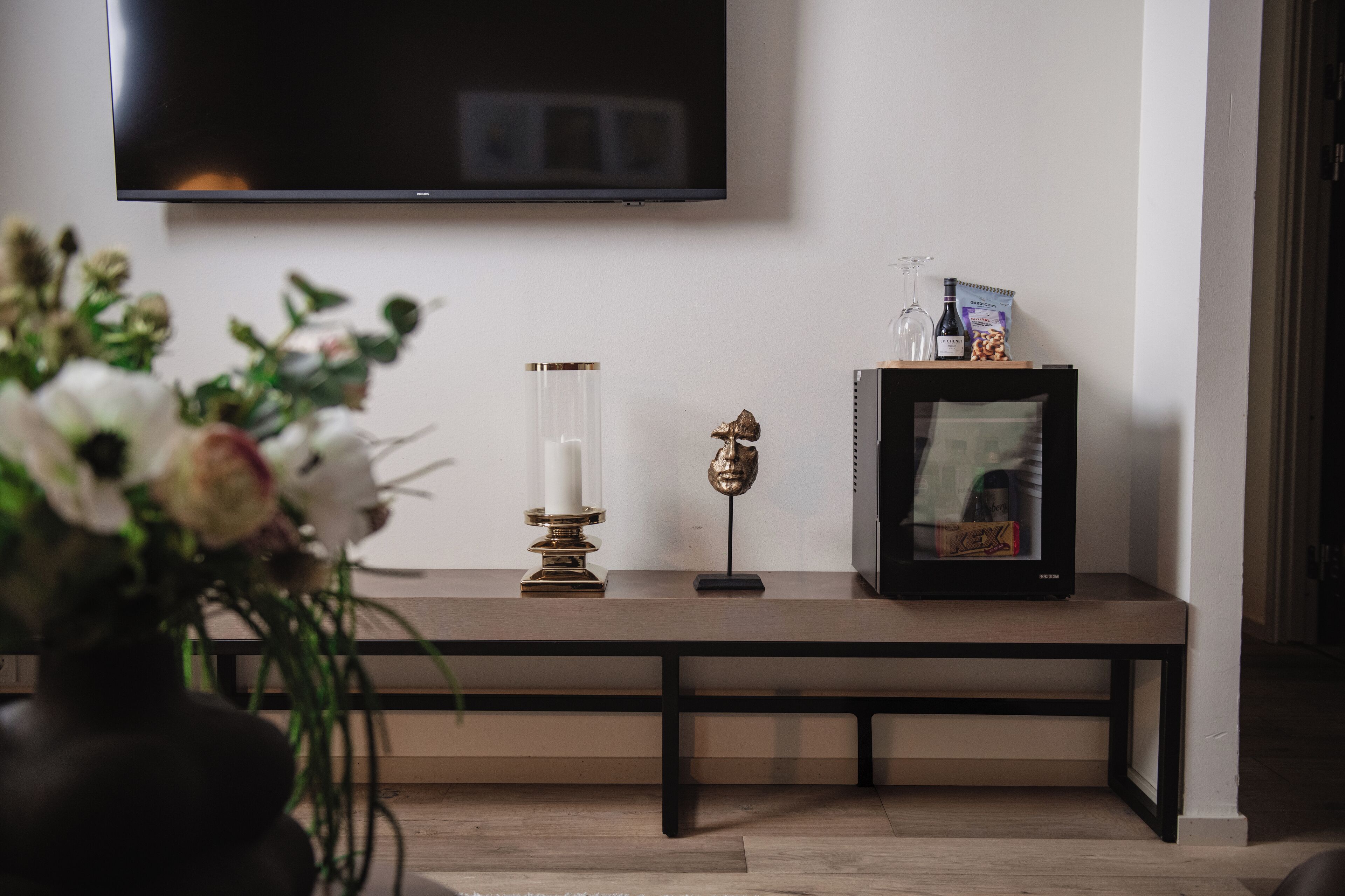A modern living room with a flat-screen TV mounted on the wall, a wooden bench with decorative items, and a vase with flowers.