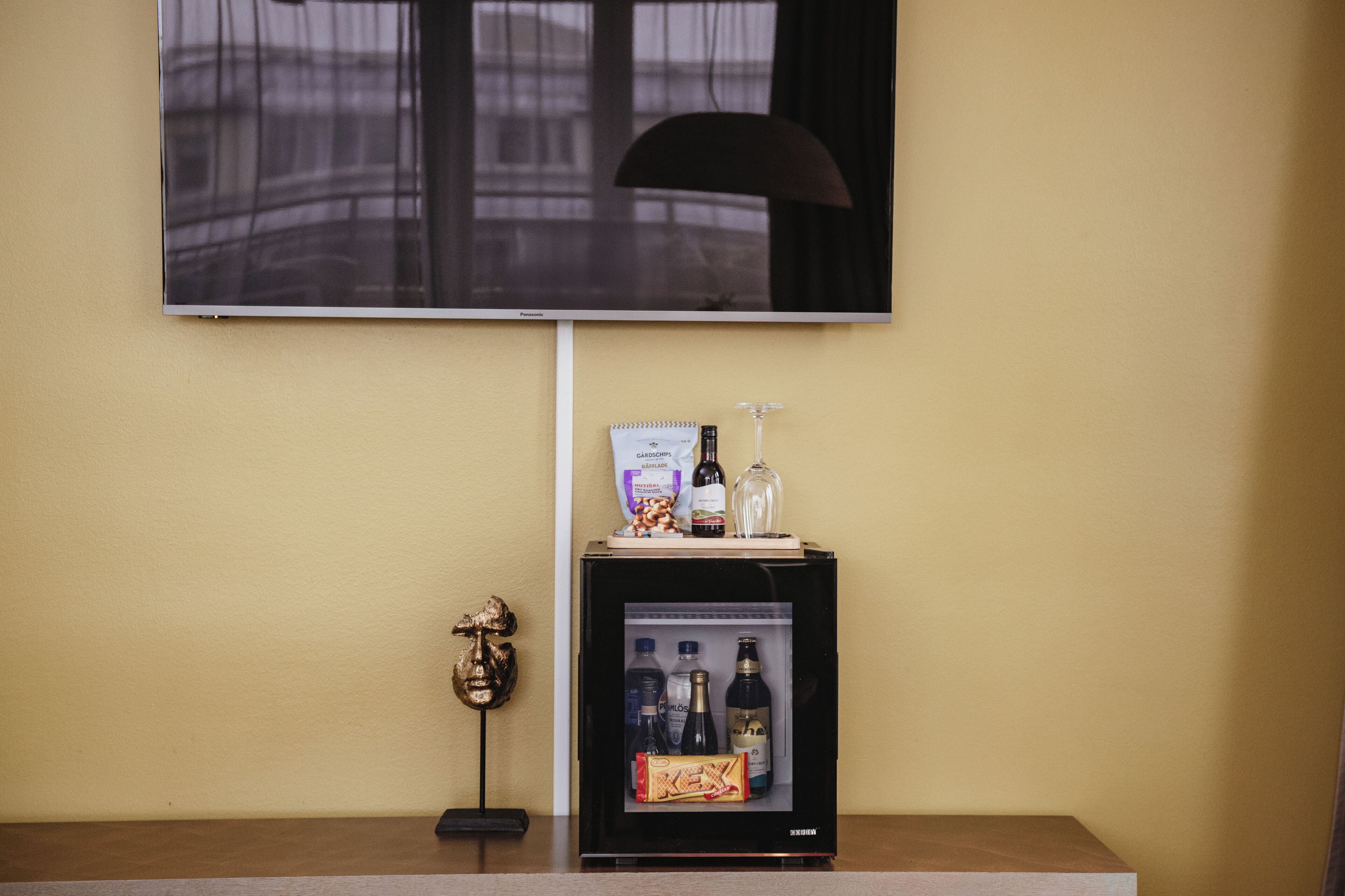 Un mini bar con un televisor encima, una pequeña estatua y un refrigerador con bebidas y aperitivos.