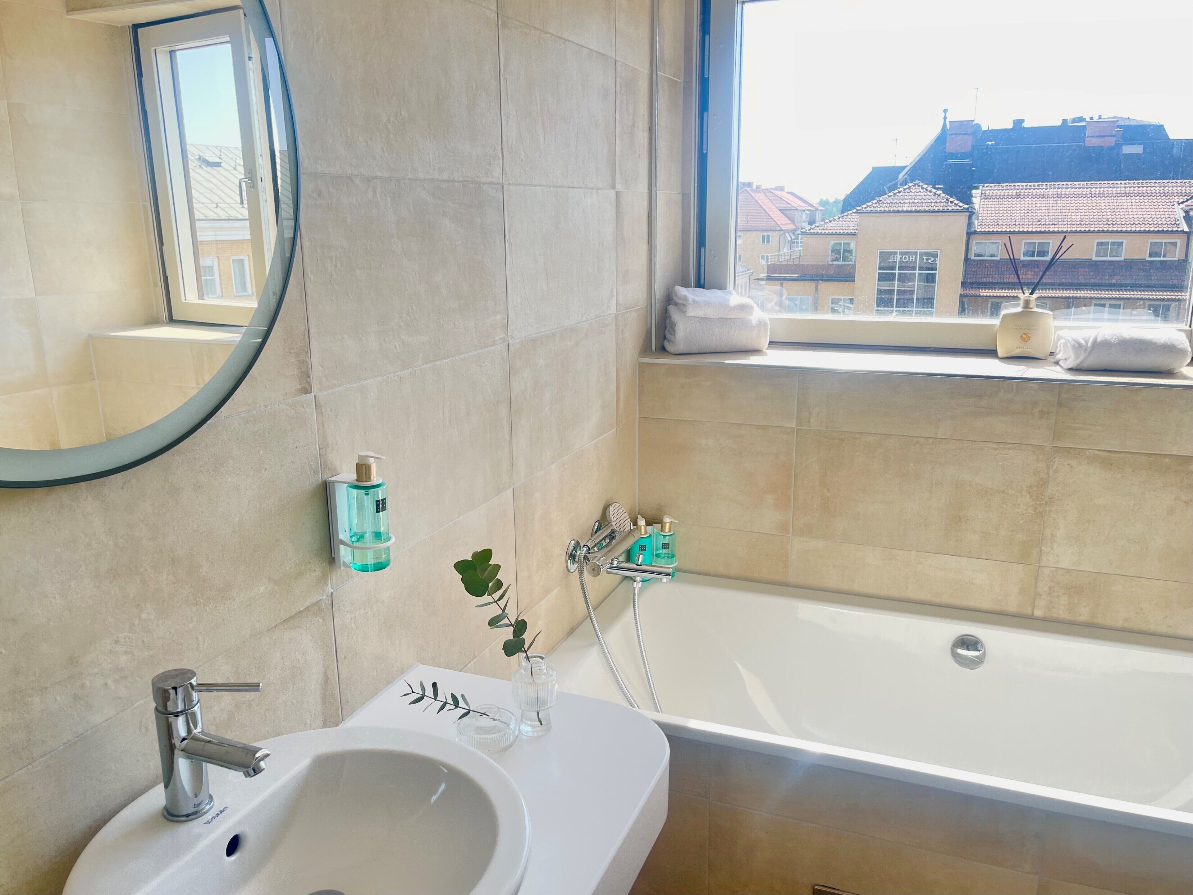 A modern bathroom with a bathtub, a sink, a mirror, and toiletries on the windowsill.