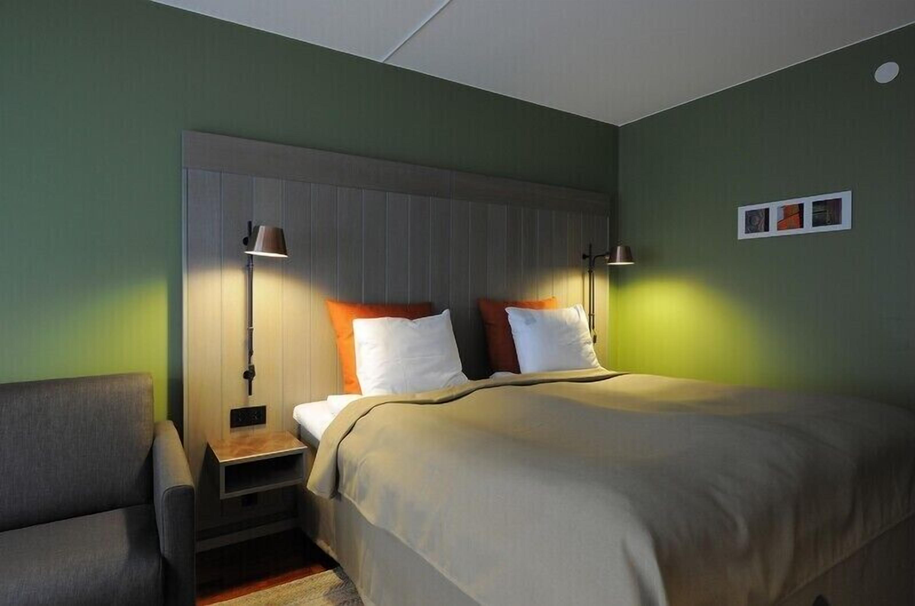 Quarto de hotel com uma cama, mesas de cabeceira, uma cadeira e uma prateleira na parede.