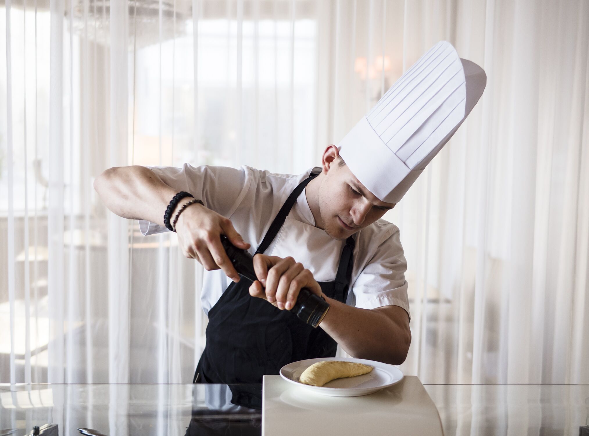 A chef in a white chef's hat and black apron preparing food in a kitchen with a glass table and white curtains in the background.