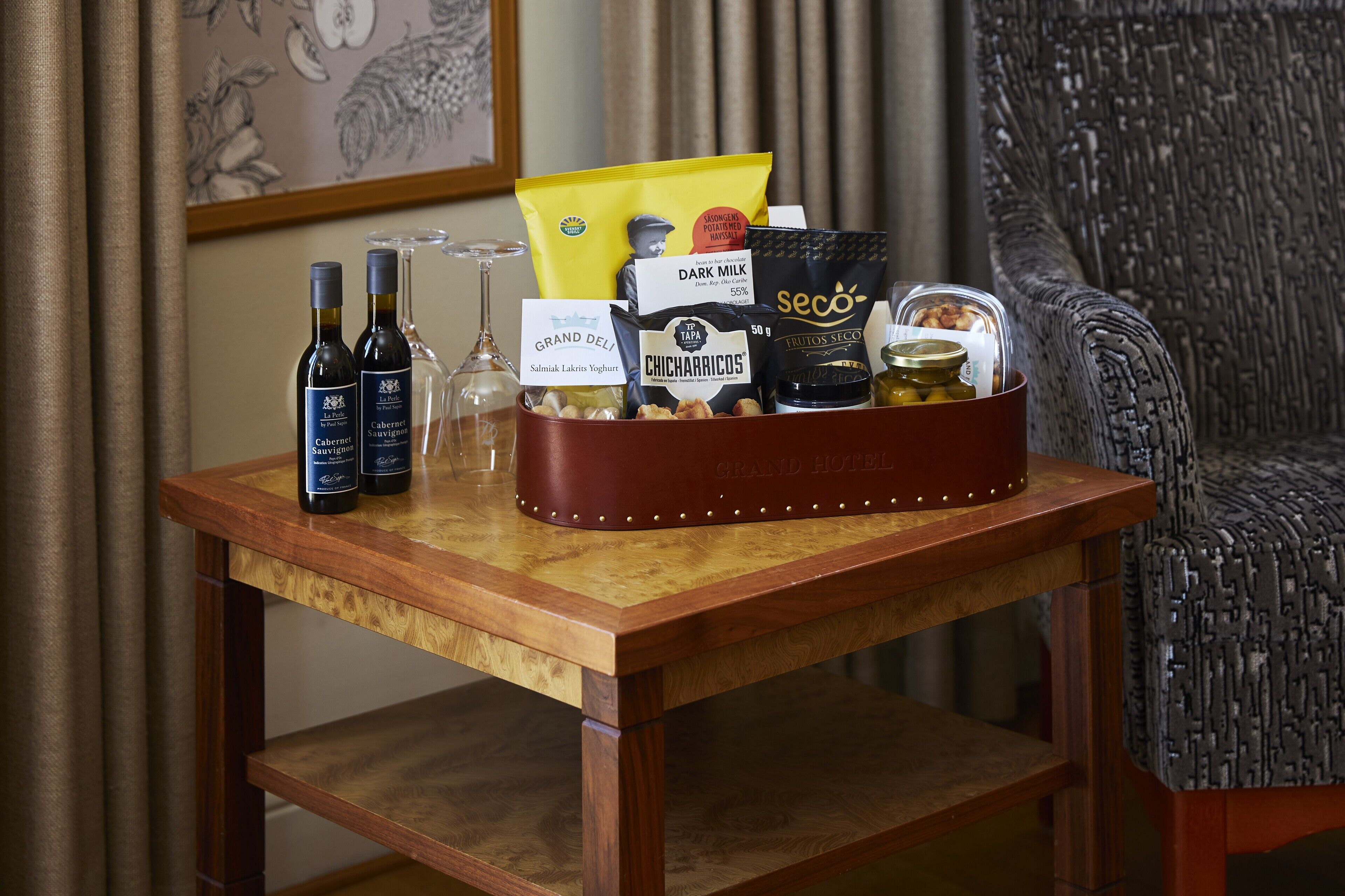 Una mesa de madera con una cesta de regalo que contiene varios artículos como botellas de vino, una copa y productos alimenticios.