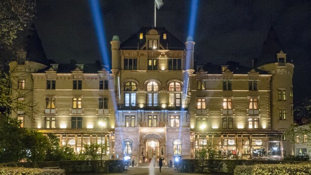 Un edificio grande e histórico con una entrada central, una fuente en primer plano y dos haces de luz destacados.