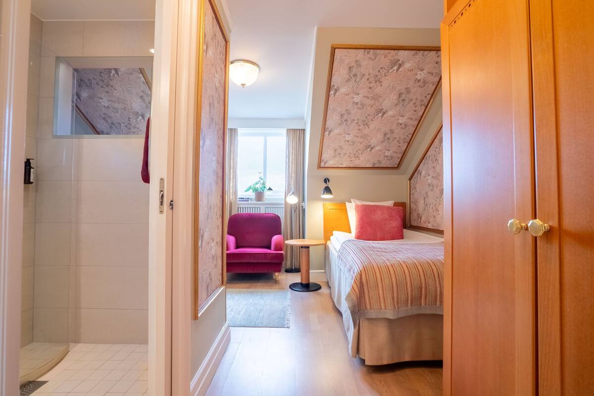 Una habitación de hotel con ducha, una cama con cabecera, una mesita pequeña y una silla.