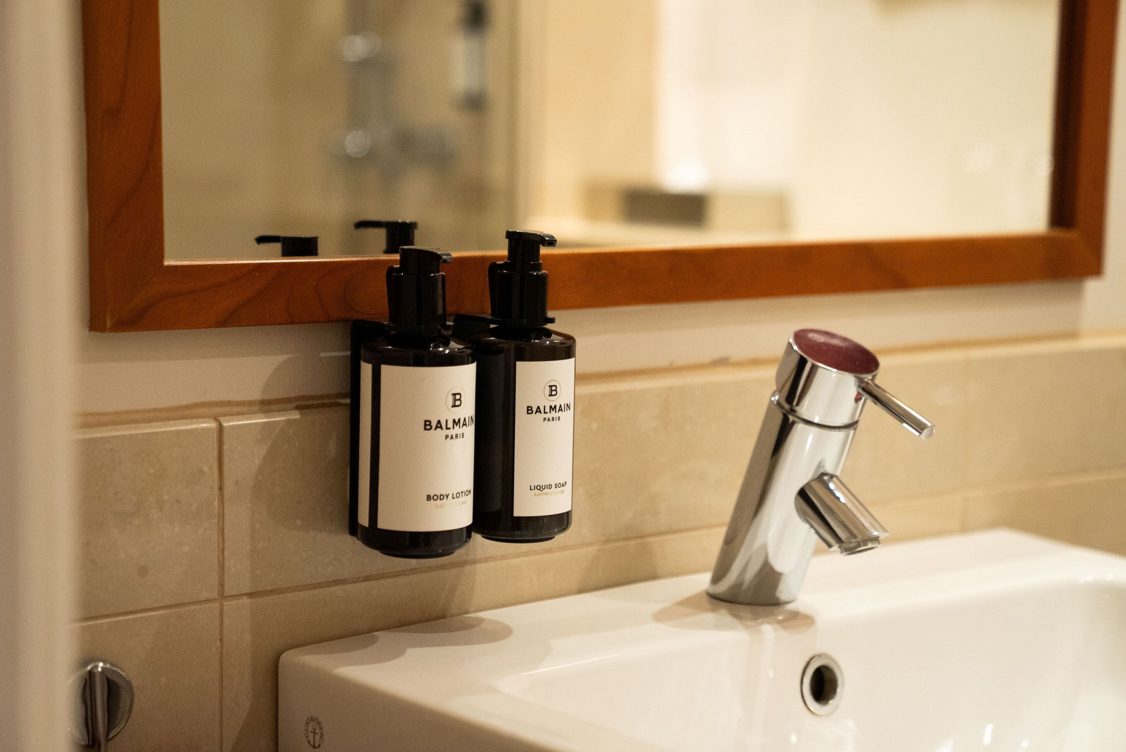 A bathroom with a white sink, a mirror with a wooden frame, and two bottles of Balmain body lotion and liquid soap.