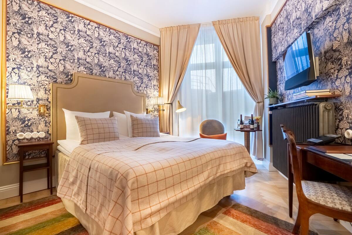 Habitación de hotel con una cama grande, un escritorio, una silla, un televisor y un ventanal con cortinas.