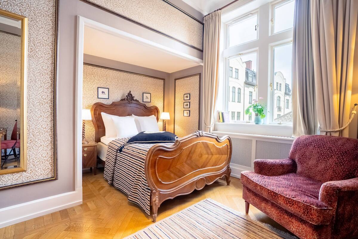 Habitación de hotel con cama, mesita de noche, silla, ventana con vistas a edificios y espejo.
