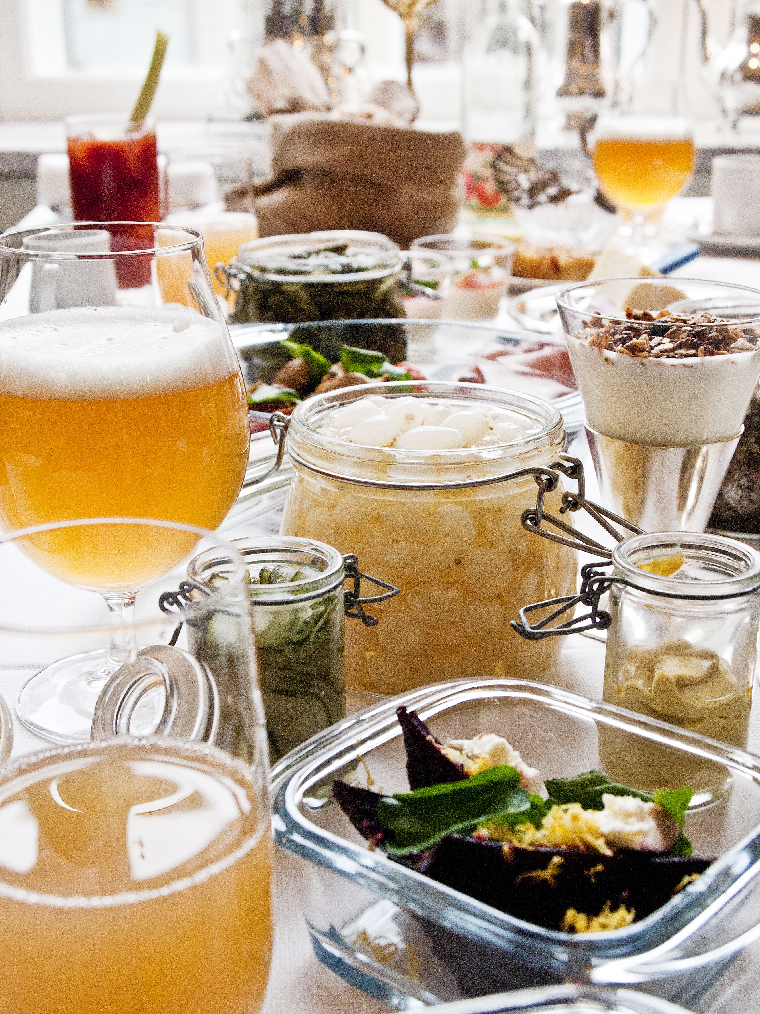 Variedad de bebidas y alimentos en una mesa, incluyendo una jarra de cerveza, un frasco de pepinillos y un plato con verduras y queso.