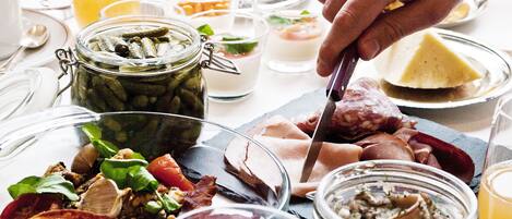Una mesa dispuesta con diversos alimentos, incluyendo frascos de pepinillos y carnes, y una persona cortando una porción de carne.
