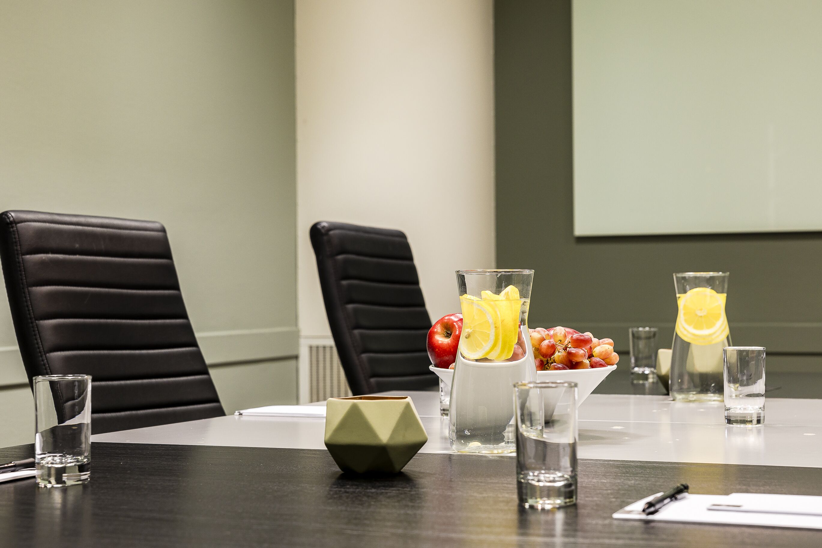 Une table de conférence avec deux chaises en cuir noir, un vase blanc contenant des fruits et des tranches de citron, et plusieurs verres d’eau.