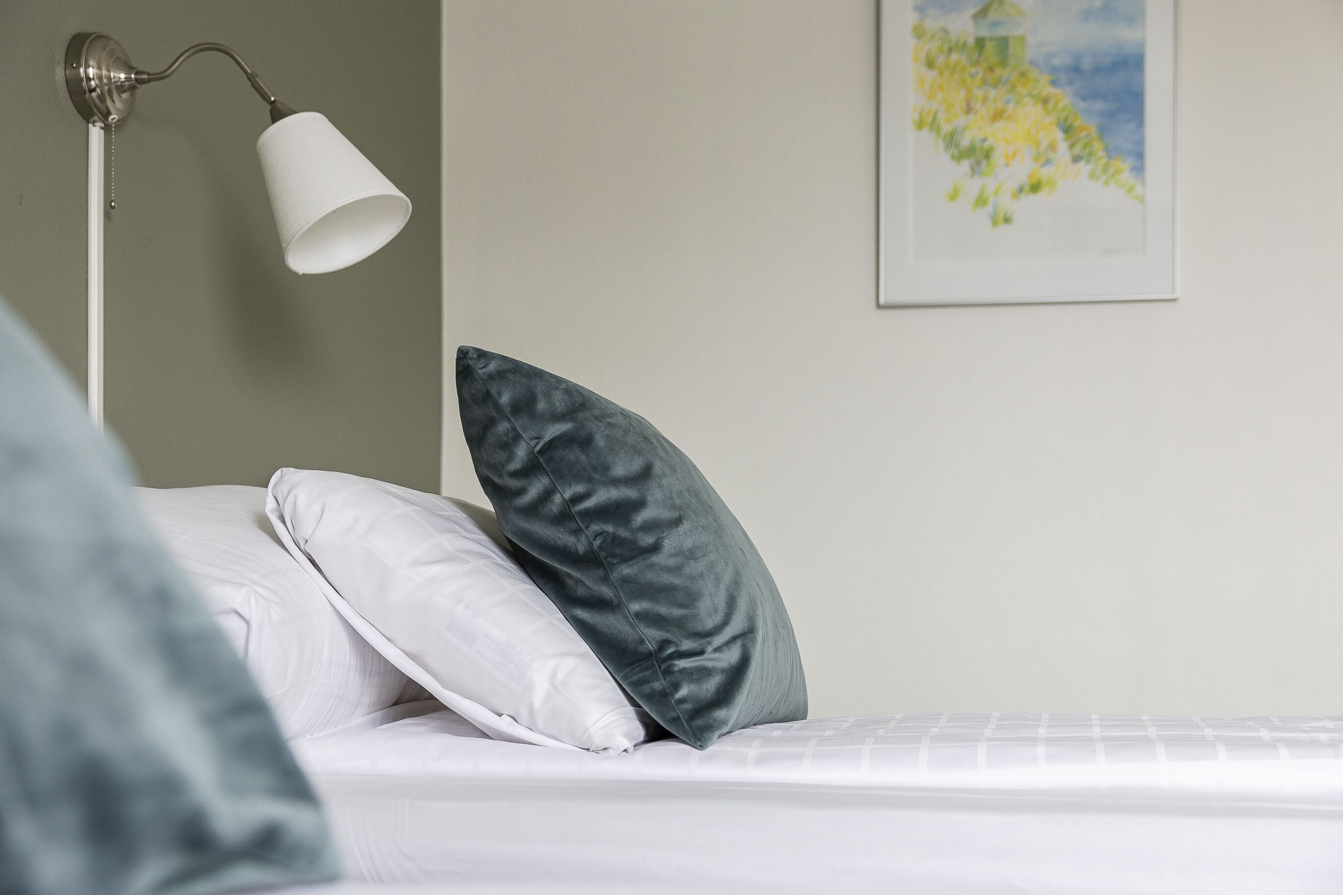 Una cama bien hecha con almohadas blancas y grises, una lámpara de pie blanca y un cuadro enmarcado con un paisaje en la pared.