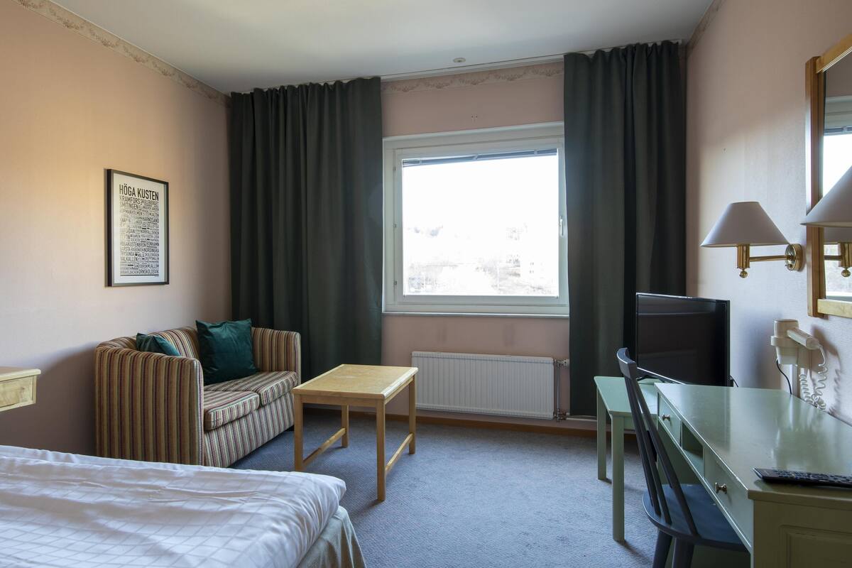 Habitación de hotel con cama, sofá a rayas, una mesita, escritorio con silla, televisión y ventana con cortinas.