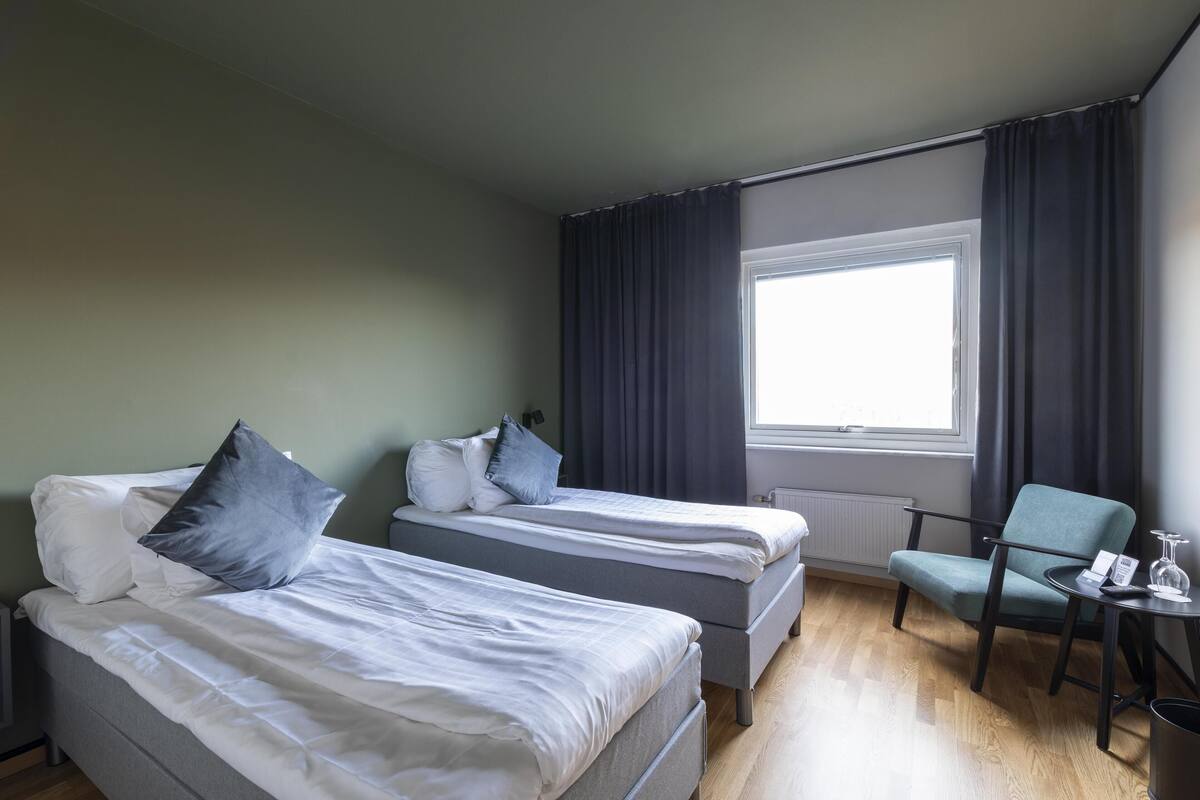 Habitación de hotel con dos camas individuales, una silla, una mesita y una ventana con cortinas.