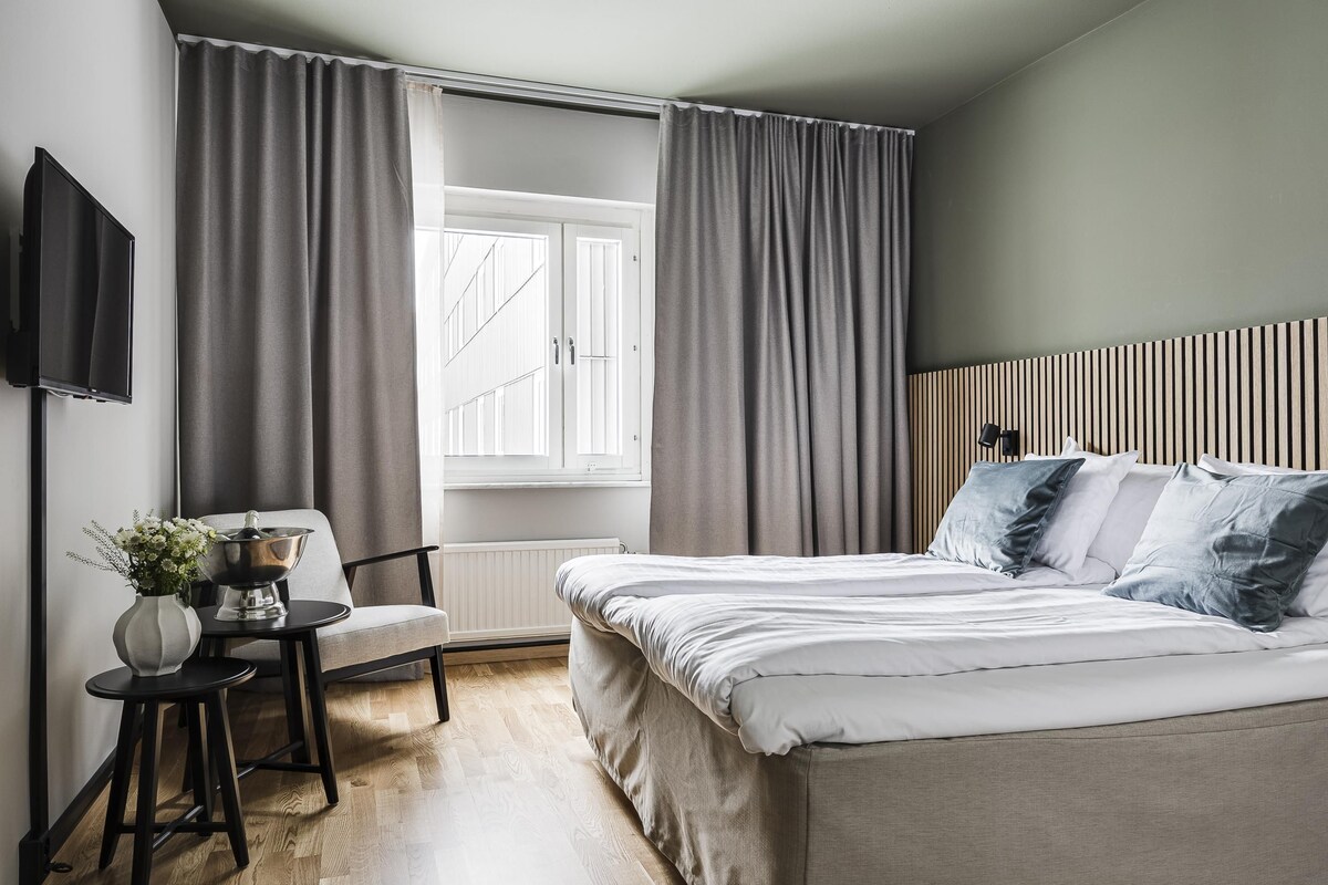 Um quarto com uma cama, uma cadeira, uma mesinha com um vaso e uma janela com cortinas.