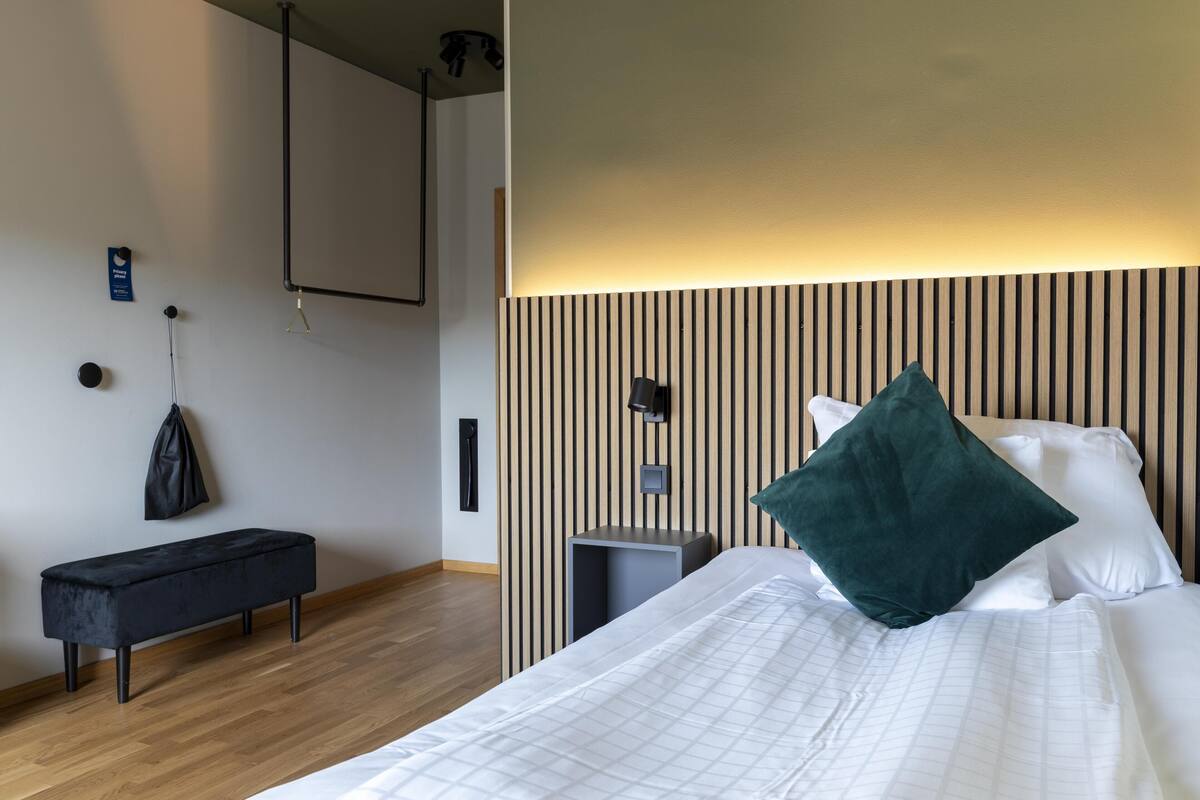 Habitación de hotel con cama, reposabajares, televisor montado en la pared y mesita de noche.