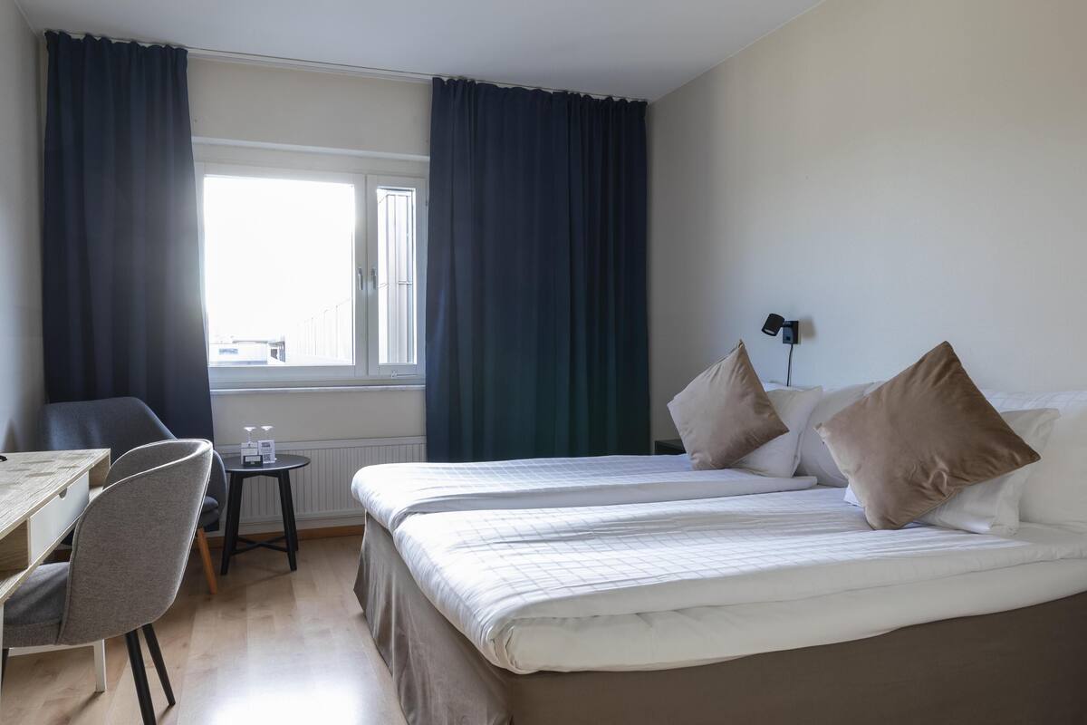 Habitación de hotel con dos camas, un escritorio, una silla y un ventanal con cortinas.