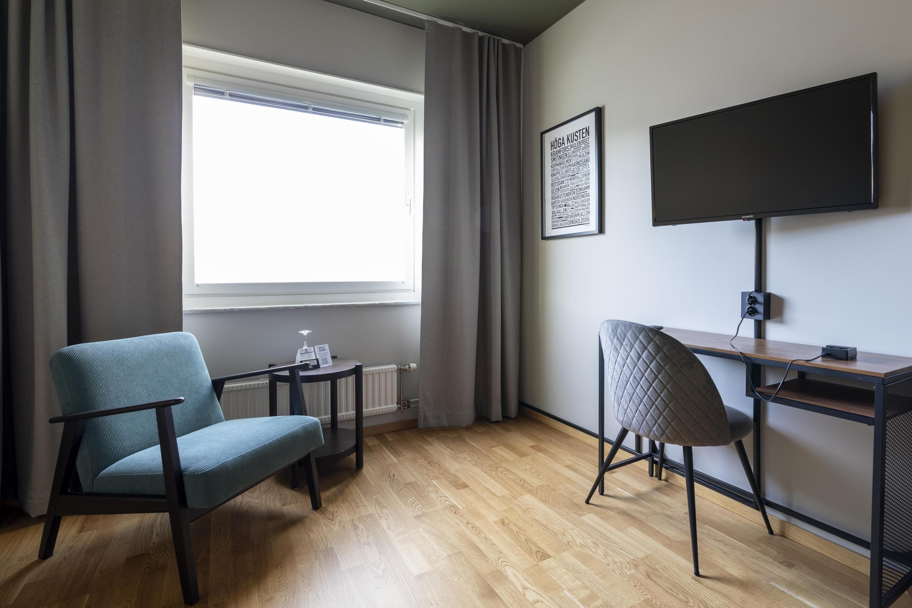 A room with a wooden floor, a desk, a chair, a window with curtains, and a flat-screen TV mounted on the wall.