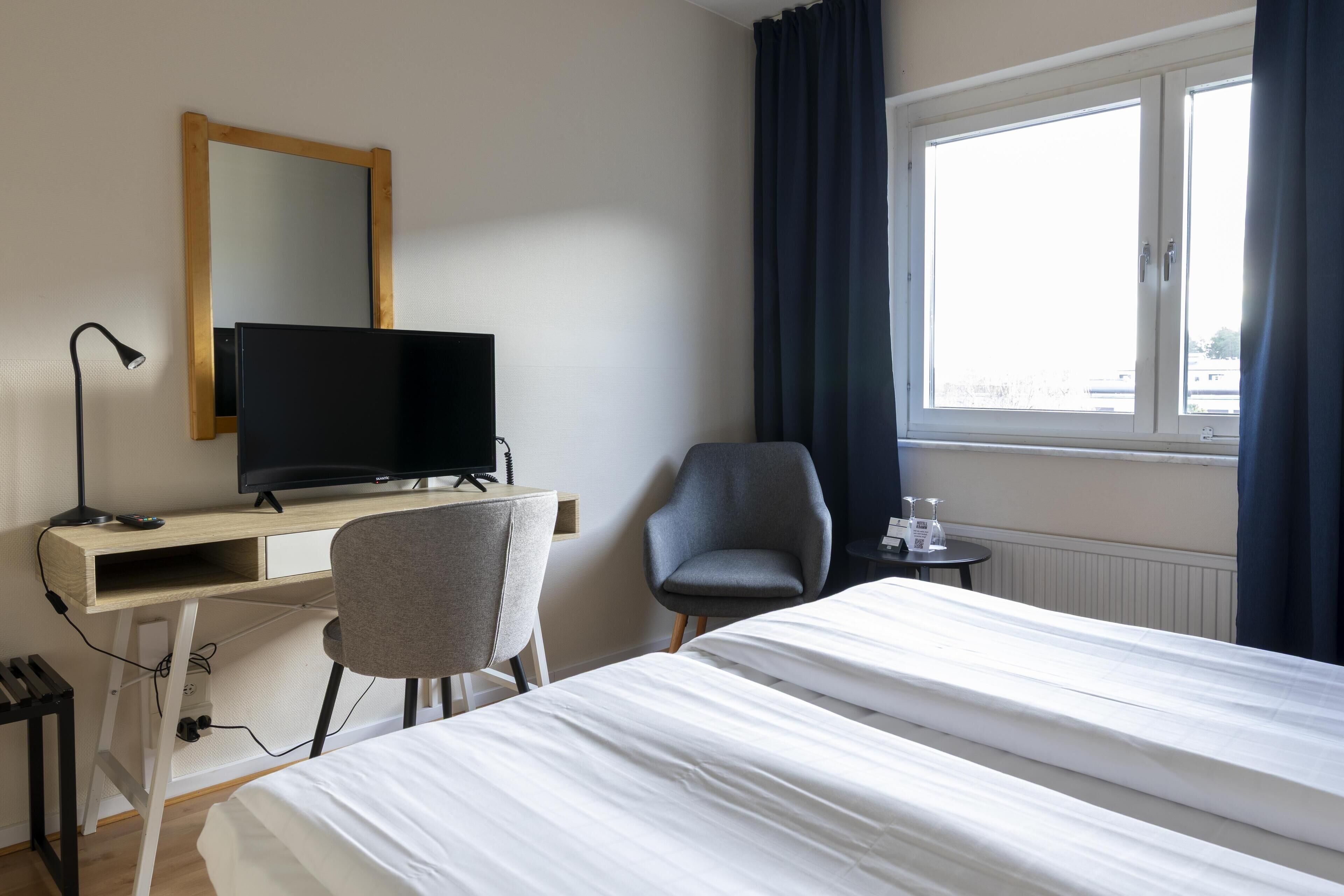 A hotel room with a bed, a desk with a computer, a chair, a TV, and a window with curtains.