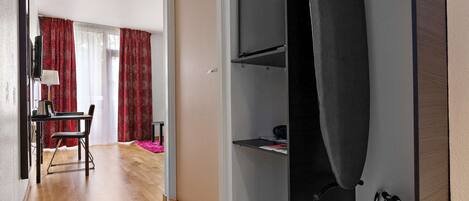 Habitación de hotel con piso de madera, un escritorio con silla, un televisor y un armario con ropa colgada.