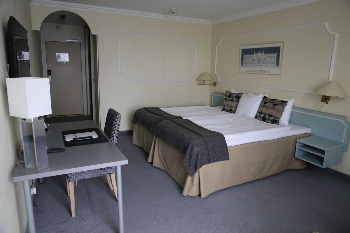 A hotel room with a bed, desk, chair, lamp, and a framed picture on the wall.