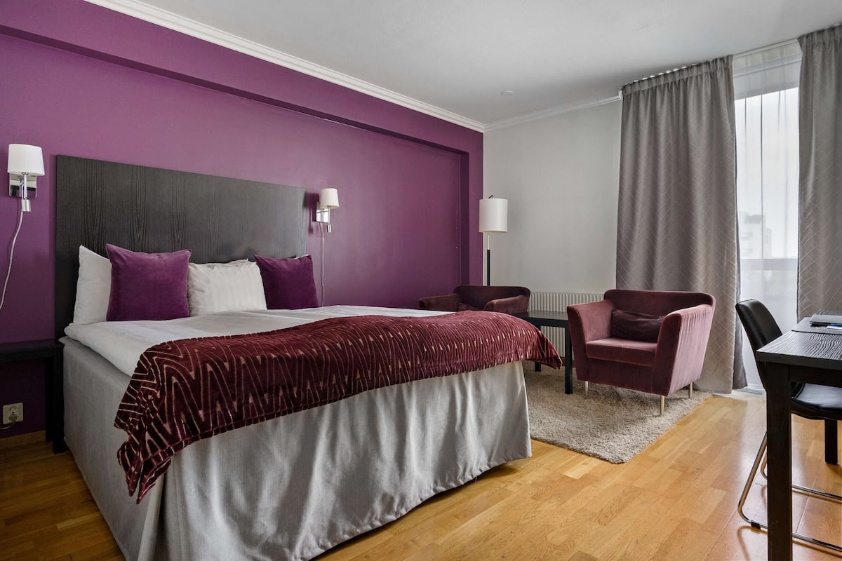 A hotel room with a large bed, a desk, a chair, and a view of the city through the window.