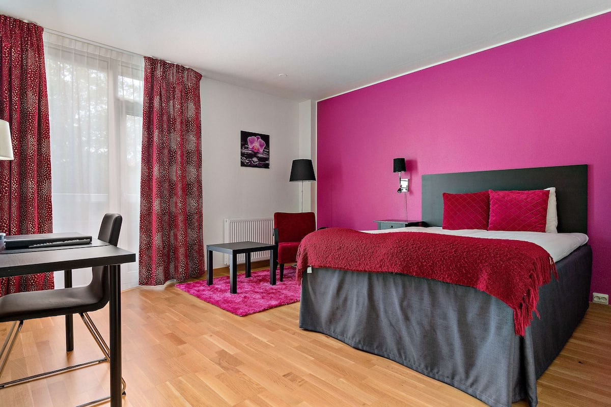 A hotel room with a bed, a desk, a chair, a radiator, and a window with curtains.