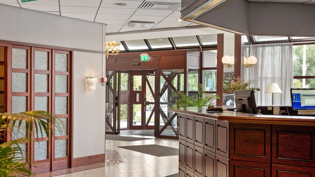 A reception area with a wooden counter, a computer, and a plant. - Best Western Royal Star (Älvsjö)