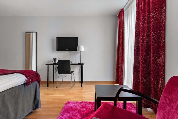 A hotel room with a bed, desk, chair, TV, and red curtains.