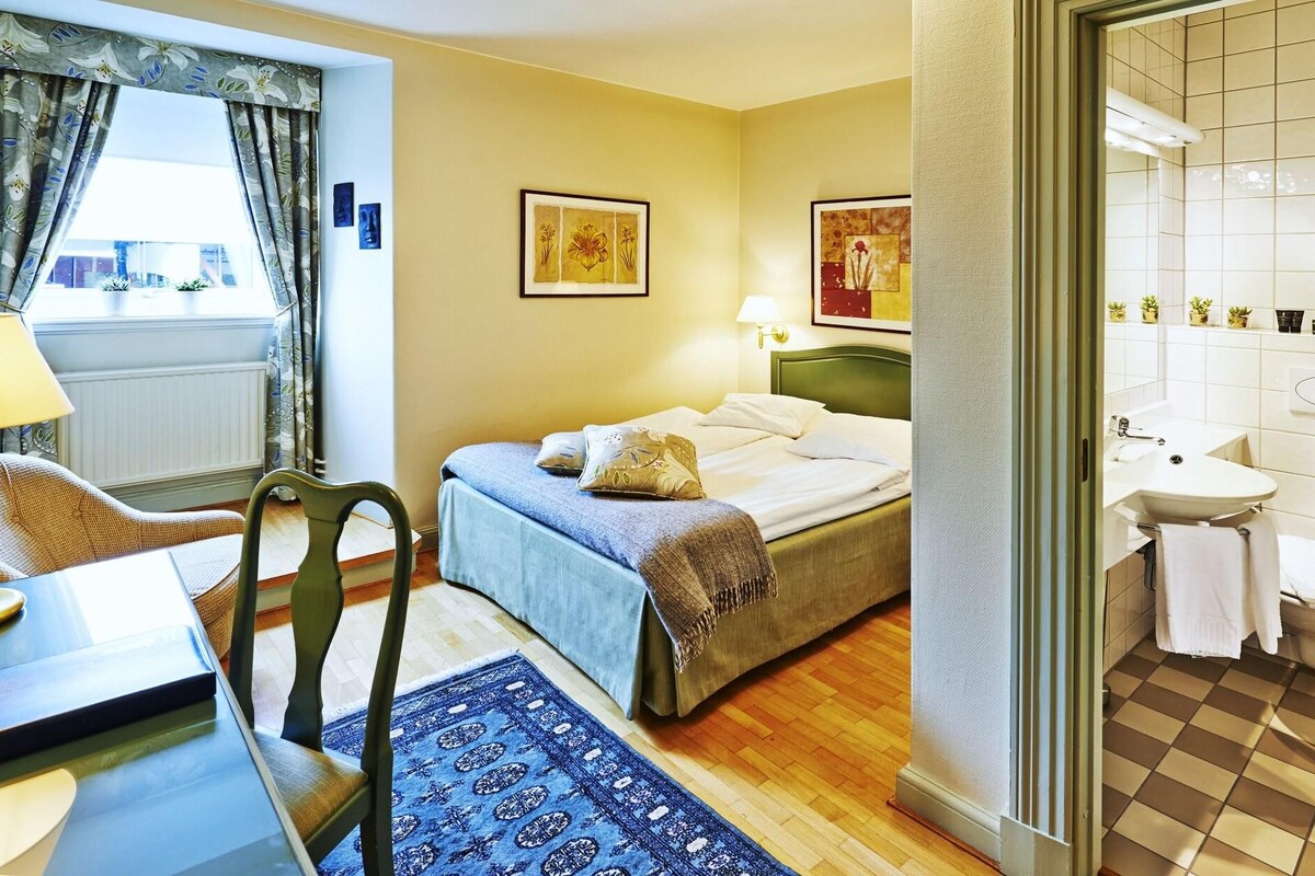 Une chambre d’hôtel comprenant un lit, une chaise, un bureau, une lampe, une fenêtre avec des rideaux, ainsi qu’une salle de bain avec un lavabo et des toilettes.