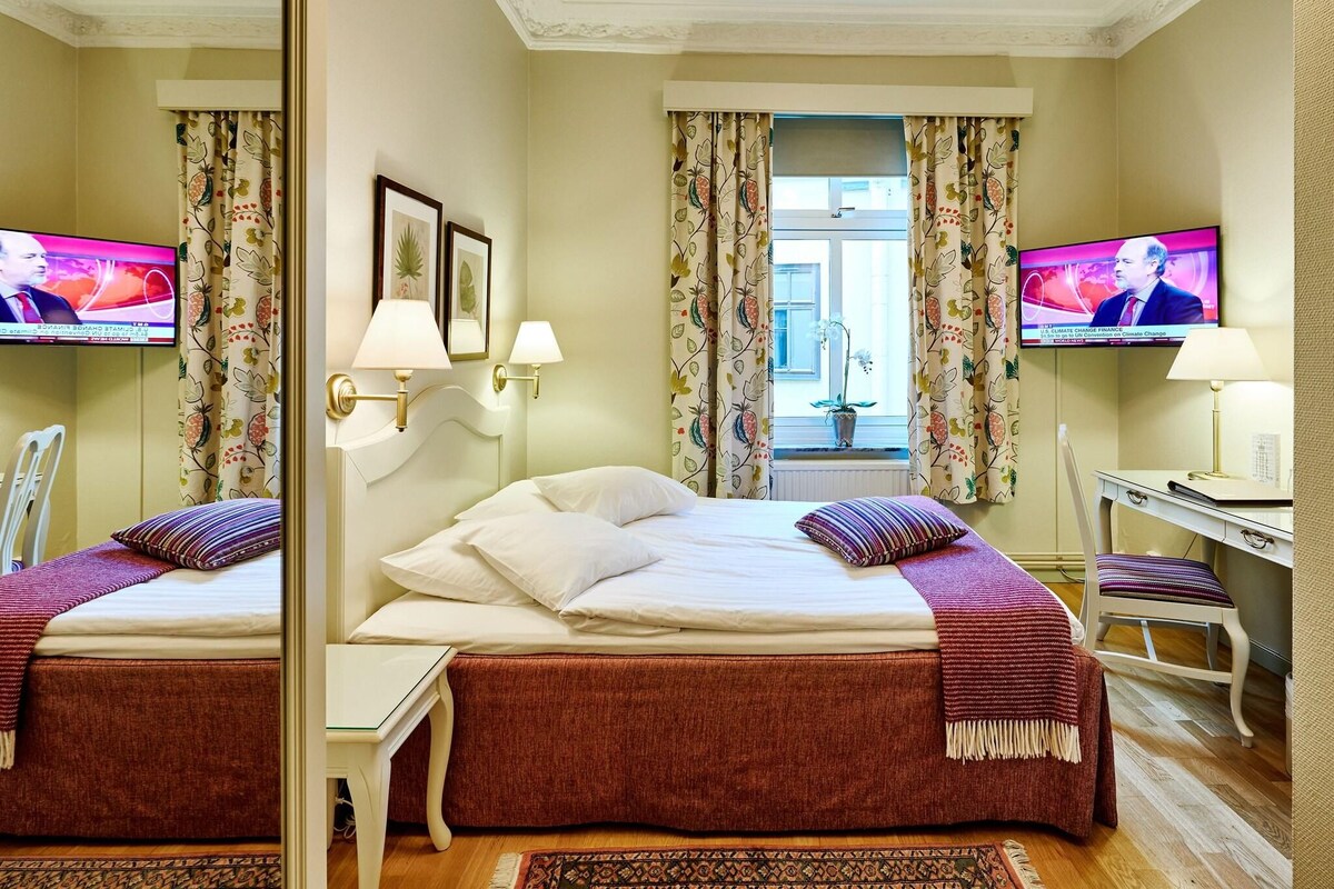 A hotel room with two beds, a desk, a chair, a TV, and a window with curtains.