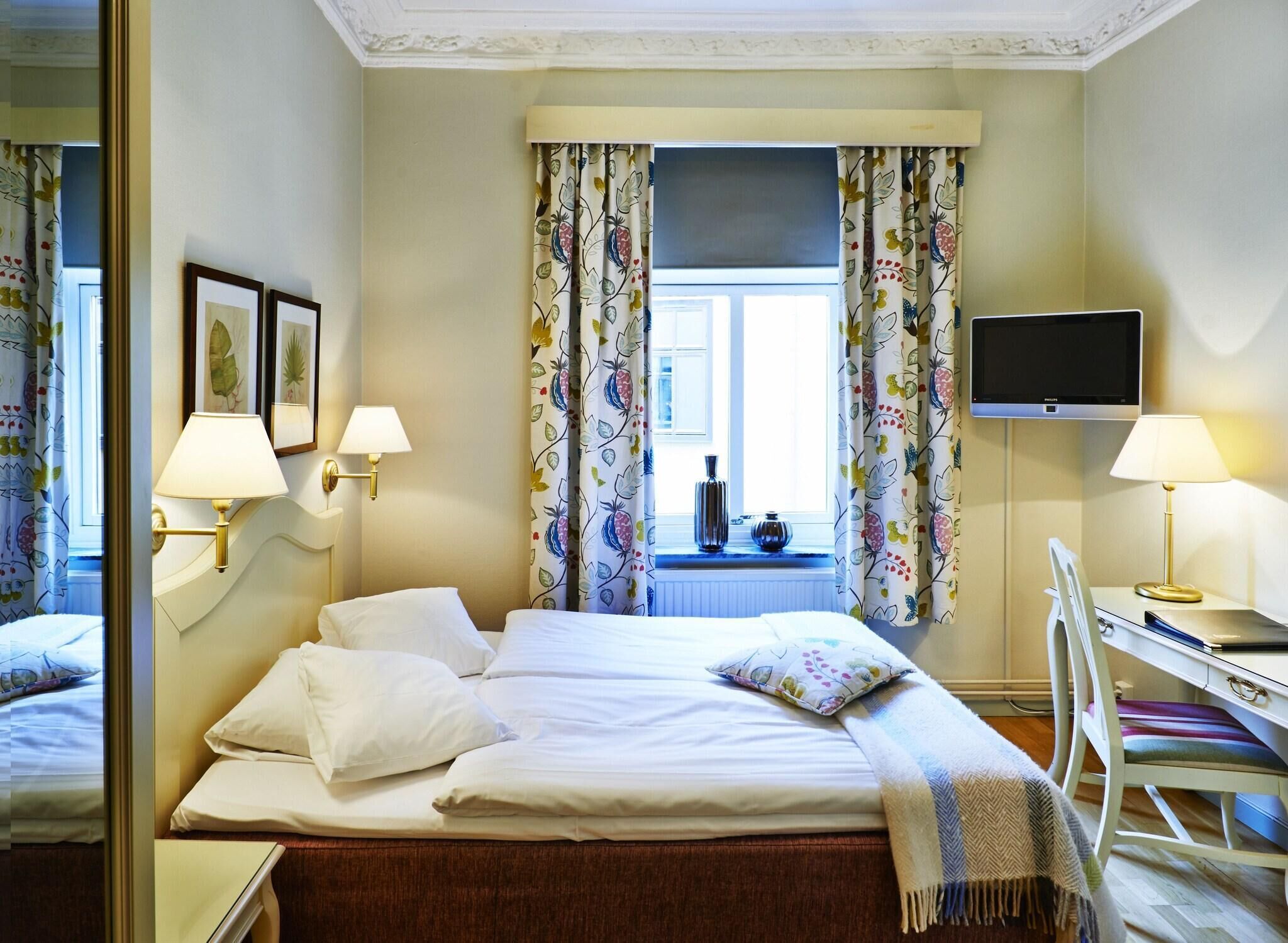 A hotel room with a bed, two bedside lamps, a TV, a desk with a chair, and a window with floral curtains.