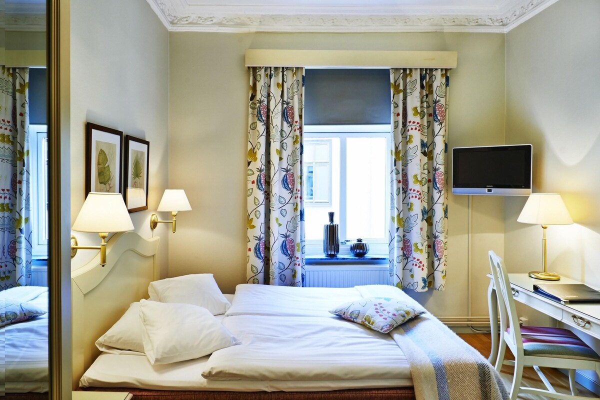A hotel room with a bed, two bedside lamps, a television, a desk with a chair, and a window with floral curtains.