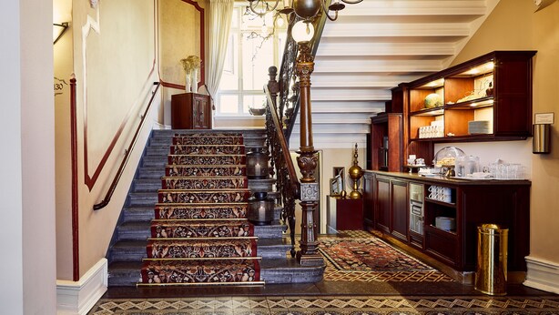 A grand staircase with ornate railings and a chandelier, leading to a room with a wooden buffet and a patterned carpet. - Hotel Royal (Gothenburg)